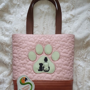 Puede incluir: Bolso tote rosa con un estampado acolchado de huellas de pata. Cuenta con una base y asas marrones. El diseño incluye una huella de pata verde con la silueta de una persona y un perro. Una etiqueta con un loro y el texto "Sunthega Designs" está adjunta.