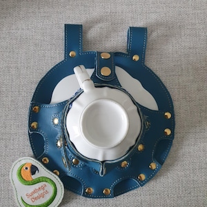Può includere: Un portateiera in pelle color teal con rivetti dorati e una tazza e un piattino bianchi. Il supporto ha due manici superiori e un design cucito decorativo. È visibile anche una piccola toppa ricamata con un disegno di pappagallo.