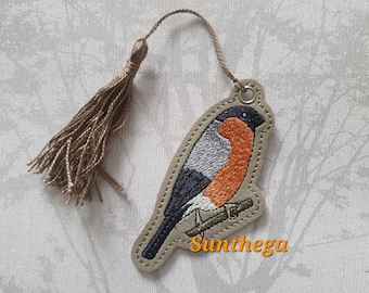 May include: A brown leather bookmark with a colorful embroidered image of a bird perched on a branch. The bookmark has a brown tassel attached to the top.