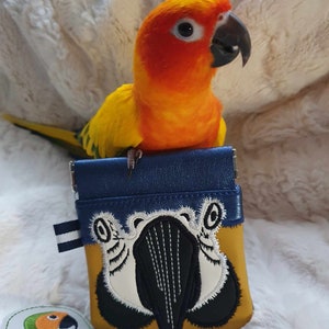 May include: A yellow and orange sun conure parrot perched on a small blue and yellow pouch with an embroidered parrot design. The pouch has a blue flap with a white stripe and a white and black embroidered parrot face on the front. The pouch is sitting on a white fluffy surface. The text "Sunthaega Designs" is on a green and orange patch in the lower left corner of the image.