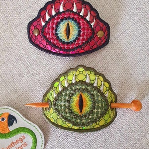 May include: Two embroidered eye hair clips, one red and one green, with a wooden hair stick. The hair clips are shaped like a dragon eye with white teeth and a yellow iris.