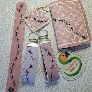 May include: A set of pink and lavender accessories featuring embroidered black ants, including a snap bracelet, a square tag, a wallet, two keychains, and a patch with a parrot graphic and the text Sunthega Designs in orange and green.
