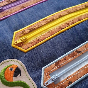 Op de afbeelding: Een close-up van een denim stof met vier verschillende gekleurde ritsen. Elke rits is genaaid in een strook kurk stof met een contrasterende gekleurde draad. De ritsen zijn paars, geel, lichtblauw en lichtbruin. Een klein geborduurd patch met een groene slang en de tekst "Suntega Designs" bevindt zich in de linkerbenedenhoek.