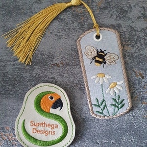 May include: A beige bookmark with a bee and daisies embroidered on it. The bookmark has a gold tassel. A beige patch with a green snake and the text "Sunthaga Designs" is also shown.