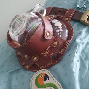 Può includere: Un porta bevande in pelle marrone con una caraffa di vetro e un bicchiere con disegni floreali. Il porta bevande è attaccato a una cintura marrone con una fibbia dorata. È visibile anche una toppa ricamata con la scritta "Sunthega Designs".
