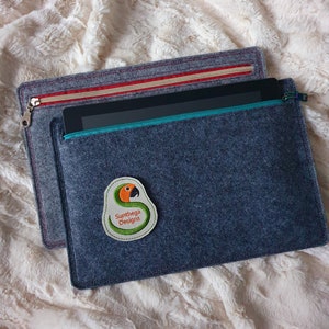 Könnte beinhalten: Eine graue Filz-Laptoptasche mit Reißverschluss. Die Tasche hat ein grünes Papageien-Logo mit dem Text "Sunthaga Designs".