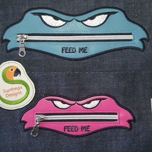 May include: Two zippered pouches in the shape of a monster's mouth, one blue and one pink, with white embroidered eyes and the text "FEED ME". A parrot logo is also visible.