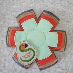 Pode incluir: Um conjunto de seis panos de cozinha de feltro em tons de cinza, marrom, laranja e verde. Os panos de cozinha têm a forma de uma estrela com um círculo no centro. Os panos de cozinha são sobrepostos. Um pequeno remendo bordado com um papagaio e o texto "Sunthaga Designs" está preso à camada inferior.