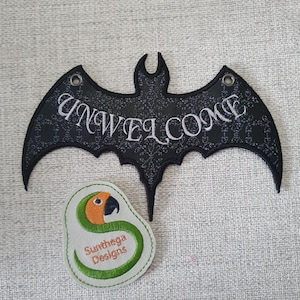 Peut inclure: Panneau noir en forme de chauve-souris avec le mot "UNWELCOME" brodé en blanc. Le panneau présente un motif décoratif et deux œillets métalliques. En dessous, un écusson avec un motif de perroquet et le texte "Sunthega Designs".