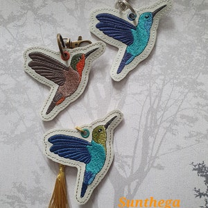 Puede incluir: Tres llaveros bordados con forma de colibrí en tonos azules, verdes, marrones y rojos. Cada colibrí está delineado con una costura en contraste y tiene un cierre metálico o borla. La palabra "Sunthega" está impresa en naranja.