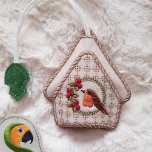 May include: A beige felt ornament shaped like a birdhouse with a red robin embroidered inside. The birdhouse has a white loop for hanging and a green felt leaf attached to the loop.