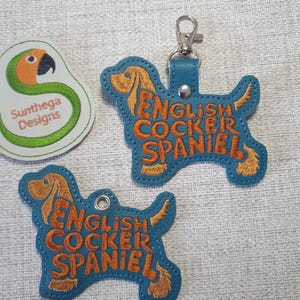 Könnte beinhalten: Ein Schlüsselanhänger und zwei passende Hundemarken mit einem englischen Cocker Spaniel in Teal. Die Hundemarken und der Schlüsselanhänger haben ein gesticktes Design mit den Worten "English Cocker Spaniel" in Orange. Ein Logo mit einem grünen und orangefarbenen Papagei ist ebenfalls sichtbar.