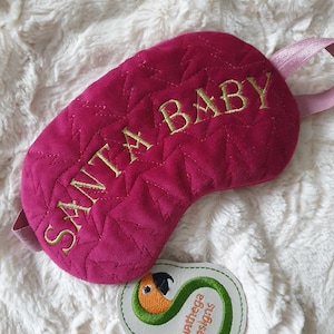 May include: A pink velvet sleep mask with gold embroidery that reads "SANTA BABY". The mask has a pink satin strap. A small white tag with a green parrot design and the text "Sunthaega Designs" is visible in the bottom right corner.