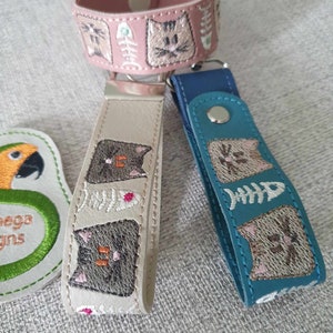 May include: Embroidered cat-themed accessories in pink, beige, and teal. The items include a pink bracelet and keychains in beige and teal. Each item features embroidered cat faces and fish bones.