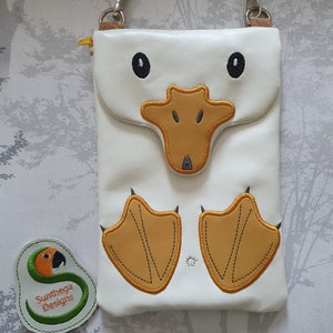 May include: White leather crossbody bag with a duck design. The bag features a flap with a duck face, black eyes, and a yellow beak. The bottom has yellow duck feet. A small patch with a parrot and the text "Sunthega Designs" is on the left.