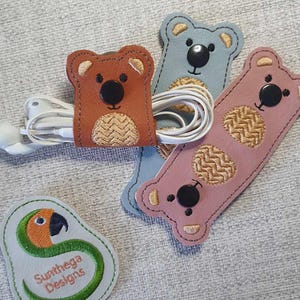 May include: Three embroidered bear-shaped cord organizers in blue, pink, and brown with black button eyes. A green and orange embroidered parrot logo is in the lower left corner. The logo reads "Sunthaga Designs".