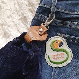 May include: A brown leather keychain with a silver clasp attached to a pair of blue jeans. The keychain has a white patch with green and orange embroidery that says "Sunthaga Designs".
