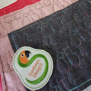 May include: A close-up of fabric pieces with embroidered cat outlines in various colors. A patch with a parrot design and the text "Sunthega Designs" is in the foreground. The fabrics are in shades of red, pink, and gray.