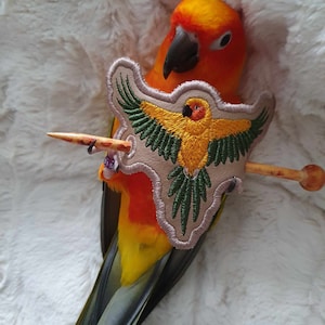 May include: A sun conure parrot with orange and yellow feathers is wearing a beige embroidered patch with a yellow bird design. The parrot is holding a wooden dowel in its beak.