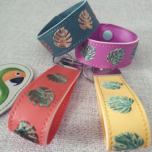 Puede incluir: Tres llaveros de cuero con diseños de hojas tropicales bordadas. Los llaveros son de color turquesa, rosa y naranja.