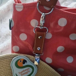 May include: A pink and white polka dot tote bag with a brown leather handle and a silver clip. The bag has a green patch with a parrot and the text "Sunthaga Designs".