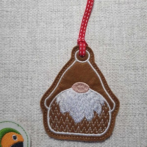 May include: A brown felt gnome ornament with a white beard and pink nose. The gnome is attached to a red ribbon with white polka dots.