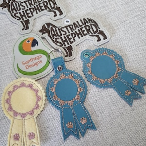 May include: A collection of embroidered keychains and patches. Includes two Australian Shepherd dog keychains, a parrot patch with text, and several blue and yellow ribbon-shaped patches with paw prints.