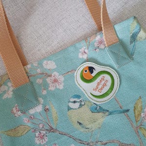 May include: A close-up of a tote bag with a floral and bird design. The bag is light blue with a pattern of pink flowers and a blue bird. It has tan straps and a patch that says "Sunthega Designs".