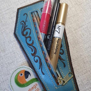 May include: A blue leather zippered pouch with a floral design and a gold zipper pull. The pouch is open and contains three makeup items: a red lipstick, a black eyeliner, and a gold tube of mascara. The pouch is sitting on a grey surface with a green and orange embroidered patch that says "Sunthega Designs".