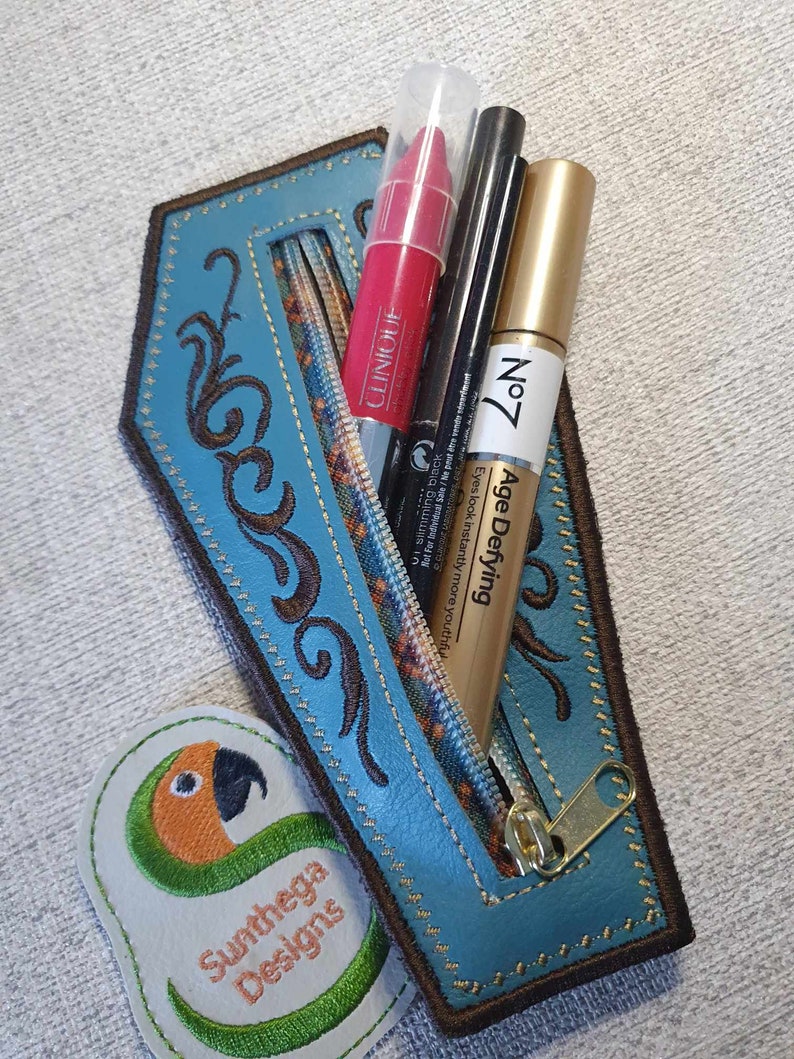 May include: A blue leather zippered pouch with a floral design and a gold zipper pull. The pouch is open and contains three makeup items: a red lipstick, a black eyeliner, and a gold tube of mascara. The pouch is sitting on a grey surface with a green and orange embroidered patch that says "Sunthega Designs".