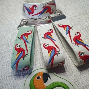 May include: A collection of parrot-themed accessories, including keychains and a wristband. The keychains and wristband feature embroidered red, blue, and yellow parrots on a light background. A parrot-shaped patch is also visible.