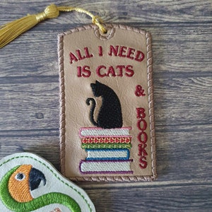 May include: A brown leather bookmark with a black cat sitting on a stack of colorful books. The bookmark has the text "All I need is cats & books" embroidered on it.