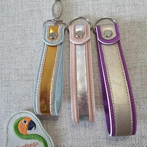 Puede incluir: Tres llaveros con correas de muñeca doradas, plateadas y moradas. Cada llavero tiene un cierre o anillo de metal. También se ve un parche verde claro con un diseño de loro y el texto "Sunthega Designs".