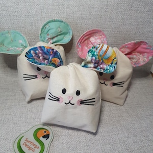 May include: Four cream-colored fabric bags with embroidered bunny faces and patterned ears. The ears feature floral and underwater designs in shades of pink, blue, and green. A small, green and white logo is in the foreground.