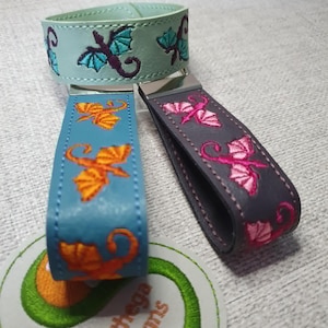 May include: Three key fobs made of leather-like material with embroidered dragon motifs. One is light green with teal dragons, one is blue with orange dragons, and one is dark grey with pink dragons. A white tag features the text Sunthega Designs.