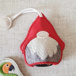Può includere: Un ornamento da gnomo in feltro rosso e bianco con una barba bianca e un pon pon bianco in cima. Lo gnomo è attaccato a uno spago bianco.