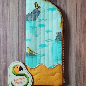 May include: A quilted pencil case with a whimsical design featuring hippos, butterflies, and fish in a water scene. The case has a teal background, a yellow base, and a zipper closure. A patch with the text "Sunthega Designs" is attached.
