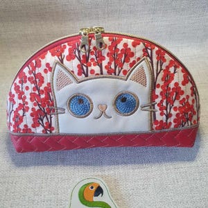 May include: A red and white quilted makeup bag with a zipper closure. The bag features an embroidered white cat with blue eyes and a pink nose. The cat is peeking out from behind a red and white floral design.