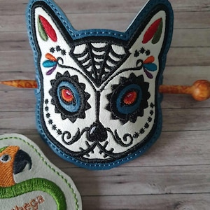 Op de afbeelding: Een blauw en wit geborduurd suiker schedel ontwerp met een spinnenweb op het voorhoofd. De schedel is bevestigd aan een houten haarstok. Het ontwerp is een populair motief voor de viering van Día de Muertos.
