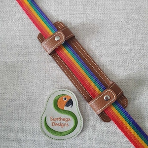May include: A rainbow striped strap with a brown leather adjustable strap. The strap has a small embroidered patch with a green snake and the text "Sunthaga Designs".