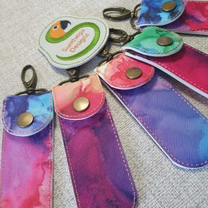 May include: A collection of keychains with colorful watercolor designs. Each keychain features a fabric case with a snap closure and a metal clasp. One keychain has a white patch with a parrot design and the text "Sumthega Designs". The color palette includes pink, purple, blue, and green.