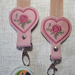 May include: Two pink heart-shaped keychains with embroidered roses and green leaves. Each keychain has a silver-colored clasp and a pink strap. A parrot-shaped keychain is visible at the bottom. The keychains are likely accessories.