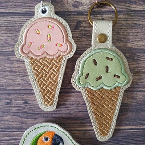 May include: Two embroidered ice cream cone keychains, one pink with sprinkles and one green with sprinkles, and a separate embroidered parrot keychain.