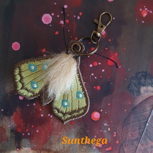May include: A green and brown embroidered butterfly keychain with a white fur accent. The keychain is attached to a bronze key ring and a clasp.