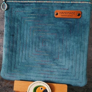 Puede incluir: Una bolsa de cremallera de terciopelo azul turquesa con un diseño acolchado que presenta un patrón cuadrado repetido. La bolsa tiene una cremallera y una etiqueta de cuero marrón que dice "HANDMADE Limited edition".