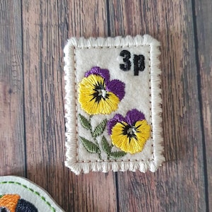 Può includere: Un cerotto di tessuto bianco con un bordo a frangia, che presenta due violette ricamate gialle e viola con steli e foglie verdi. Il cerotto ha "3p" ricamato in nero nell'angolo in alto a destra.