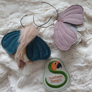 Könnte beinhalten: Zwei Filz-Schmetterlings-Spielzeuge mit Drahtantennen. Die Schmetterlinge sind türkis und lila. Ein grünes gesticktes Patch mit einem Papageien-Design und dem Text "Sunthaga Designs" befindet sich in der unteren rechten Ecke.
