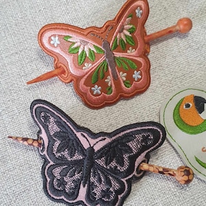 Op de afbeelding: Twee vlinder haarspeldjes met houten stokjes. De bovenste speld is oranje met groene en witte bloemenborduurwerk. De onderste speld is zwart met een roze achtergrond.