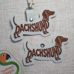May include: Two light blue dachshund keyrings with brown embroidered details. The word "DACHSHUND" is stitched across the dog's body. One keyring has a silver clasp. A green and orange parrot patch with the text "Sunthega Designs" is also visible.