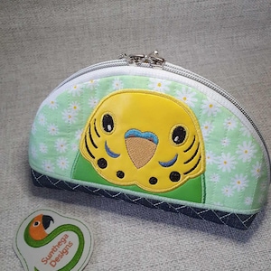 Puede incluir: Una bolsa de cremallera verde y amarilla con un patrón de margaritas y una linda cara de periquito bordada. La bolsa tiene un fondo blanco con ribete verde y un cierre de cremallera.